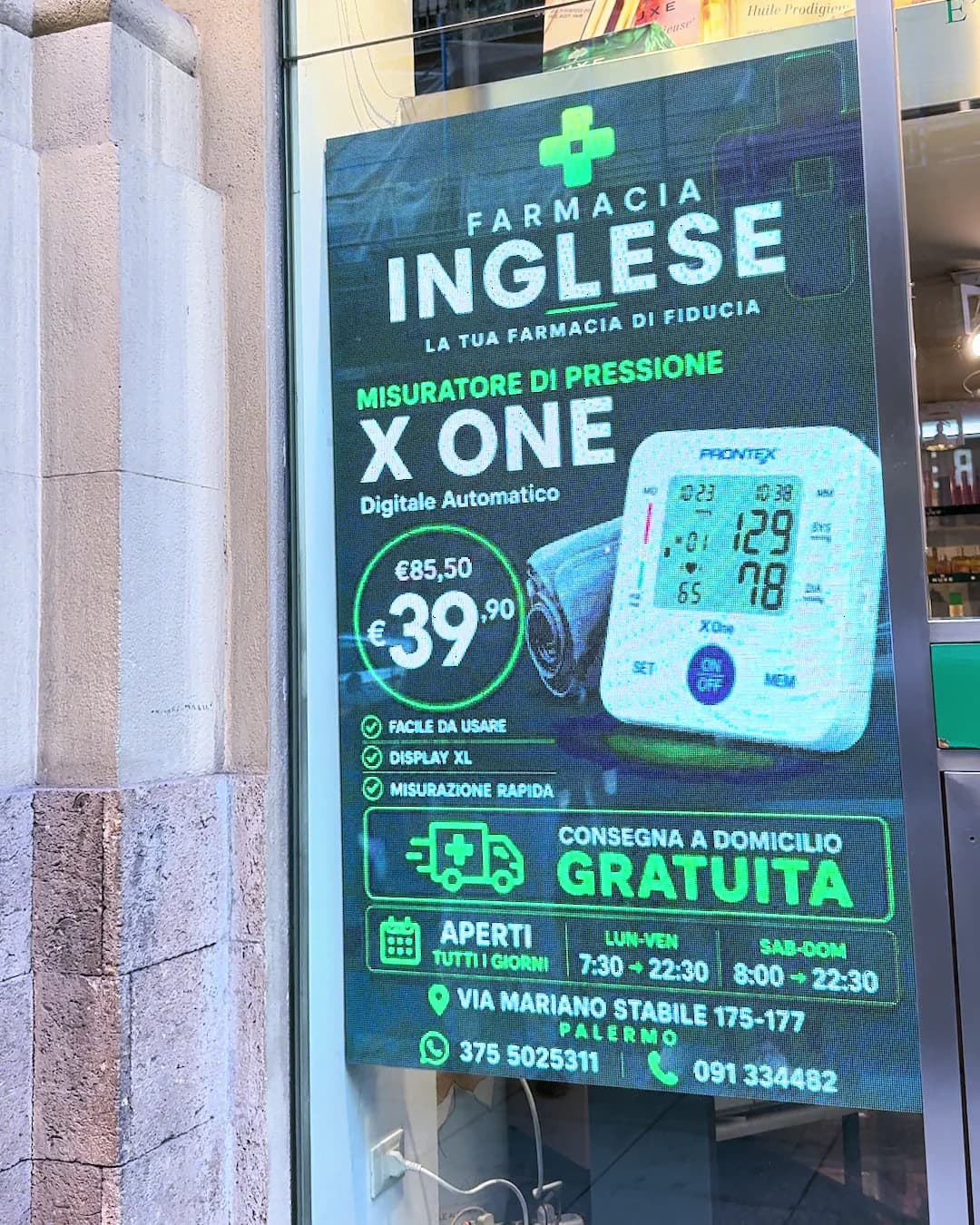 Il display in vetrina della Farmacia Inglese visto dalla strada, fra la pietra della facciata e il riflesso delle insegne.