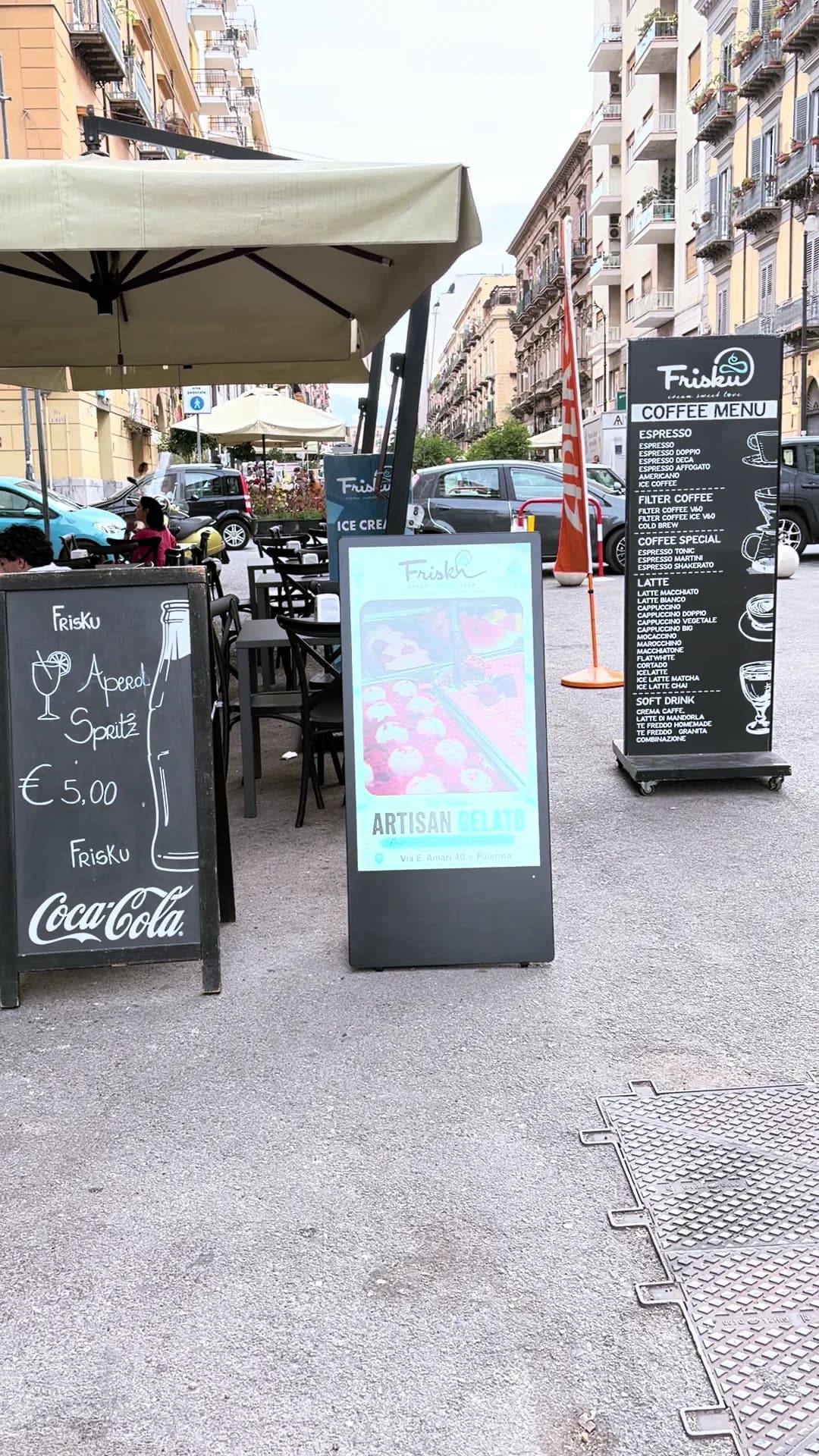 Il dehors del bar visto per intero: ombrelloni, tavoli, la fila di lavagne e il totem digitale sulla strada.