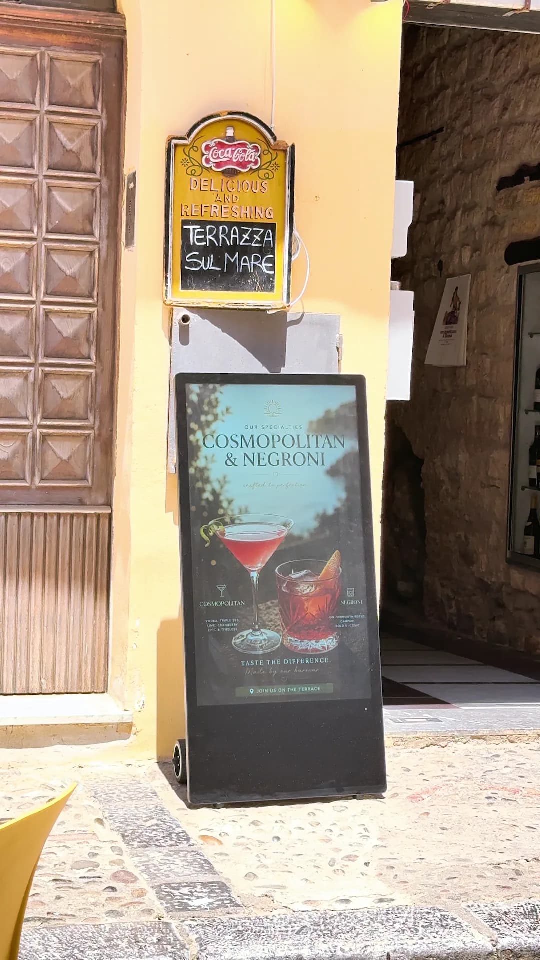 Totem digitale acceso contro un muro giallo al sole, con la proposta di due cocktail; sopra, l’insegna Terrazza sul Mare.