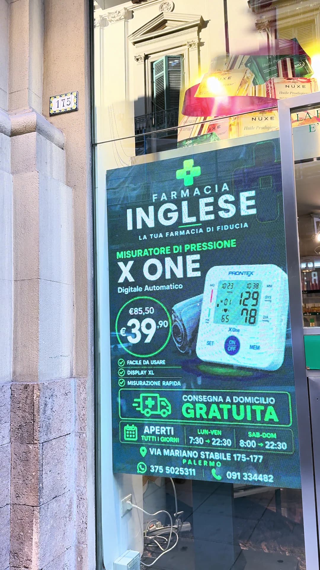 Display verticale acceso nella vetrina della Farmacia Inglese, con la promozione di un misuratore di pressione, orari e indirizzo.