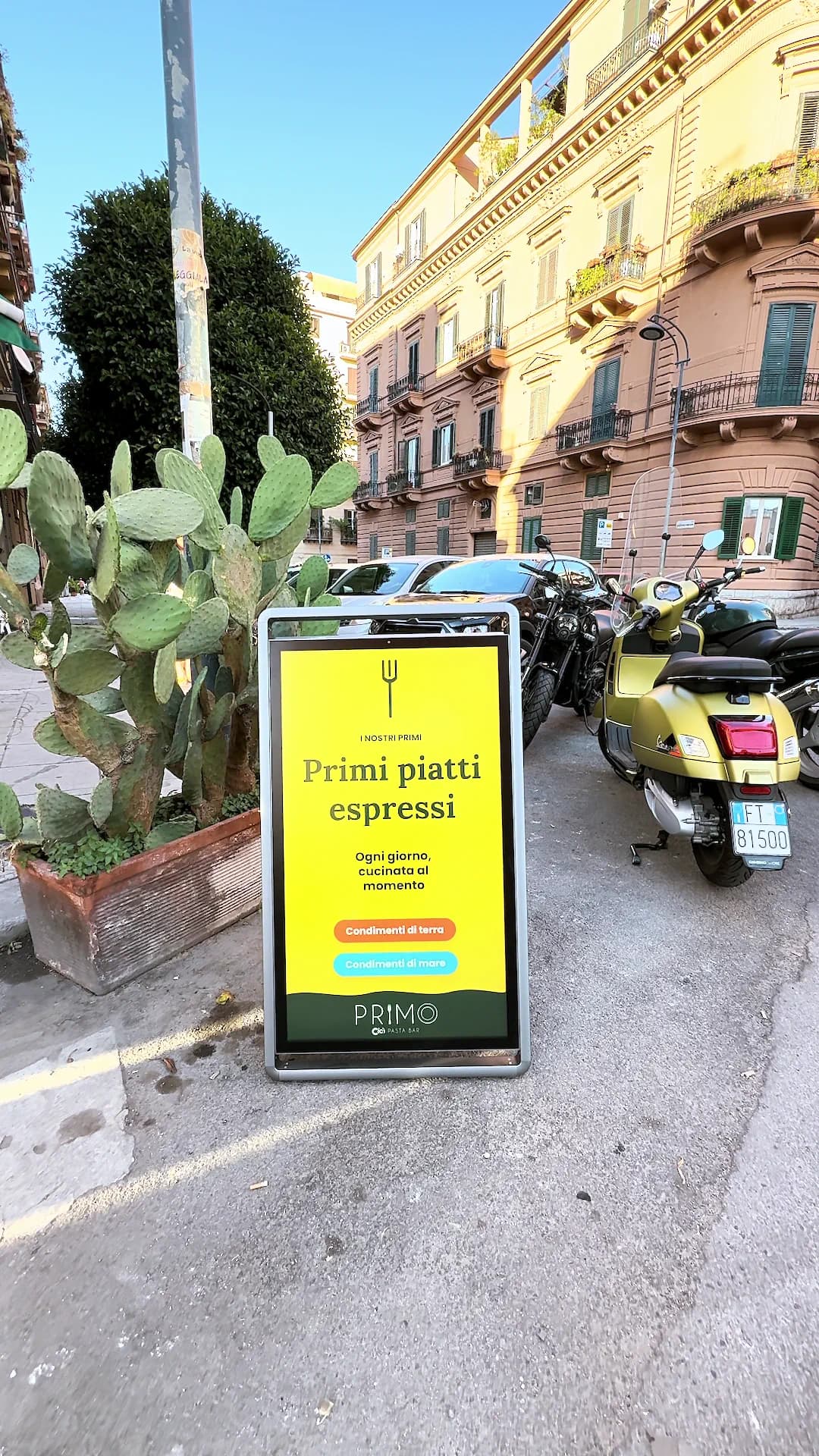 Totem digitale su un marciapiede di Palermo con la schermata gialla dei primi piatti, accanto a un fico d’India e a una fila di motorini.